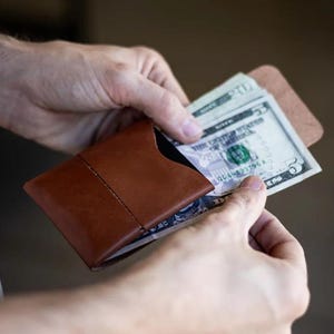 May include: A brown leather wallet with a minimalist design, held open to reveal several U.S. dollar bills. The wallet is rectangular with a stitched seam down the middle. The bills are visible, with a five-dollar bill prominently displayed.
