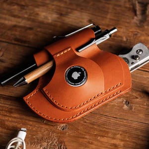 Könnte beinhalten: Ein brauner Leder-Organizer mit Stift, Messer und Apple AirTag. Der Organizer ist mit weißem Faden genäht und hat ein kreisförmiges Apple-Logo. Ein Akkordeon und ein Ladekabel sind ebenfalls sichtbar.