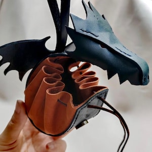 以下が含まれることがあります： 茶色と黒の色合いの、手作りの革製ドラゴンをテーマにしたポーチ。ポーチはドラゴンの頭と翼のデザインが特徴で、巾着開閉式です。内側は明るい茶色で、暗い外観とは対照的です。ユニークなファンタジーアクセサリー。
