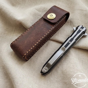 Könnte beinhalten: Eine braune Leder-Messerscheide mit Messing-Druckknopfverschluss neben einem schwarz-silbernen Klappmesser. Das Messer hat einen Taschenclip.