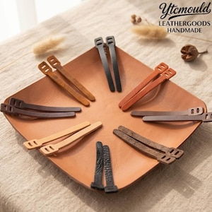 May include: A selection of leather hair clips in shades of brown and black, displayed on a square leather tray. The clips feature a straightforward, practical design. The tray rests on a neutral-toned surface. The text "Ytcmould LEATHERGOODS HANDMADE" is visible.