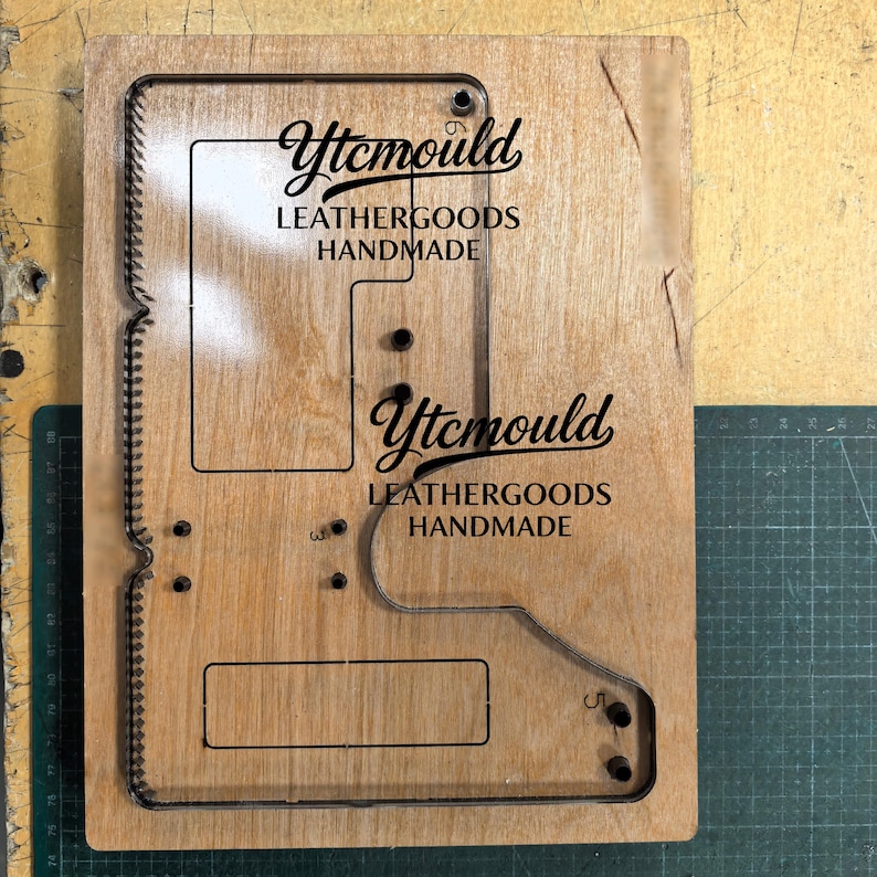 K&ouml;nnte beinhalten: Eine h&ouml;lzerne Lederhandwerks-Stanzform mit komplexem Design. Die Form weist ausgeschnittene Formen und den Text "Ytcmould LEATHERGOODS HANDMADE" in Schwarz auf. Das Holz hat eine nat&uuml;rliche Maserung und liegt auf einer gr&uuml;nen Schneidematte.