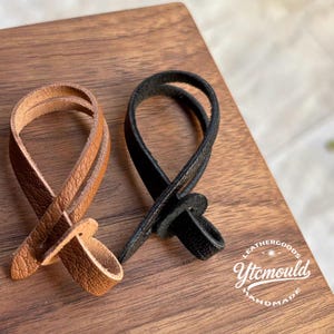 May include: Two leather loops, one tan and one black, lie on a wooden surface. The tan loop is made of layered leather strips, while the black loop appears to be a single piece. The loops are likely for crafting or leather goods.