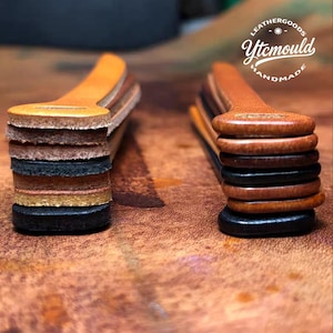 Op de afbeelding: Twee stapels leren stukken in verschillende tinten bruin, beige en zwart. Elke stapel heeft een handvat-achtige vorm aan de bovenkant. De stapels staan op een bruin, getextureerd oppervlak. De tekst "LEATHERGOODS Ytcmould HANDMADE" is zichtbaar.