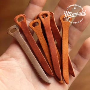 May include: A collection of leather craft tools in various shades of brown and orange. Each tool has a slender, elongated shape with a rounded top featuring a small hole. The tools are handmade, as indicated by the text "Leathergoods Ytcmould Handmade".