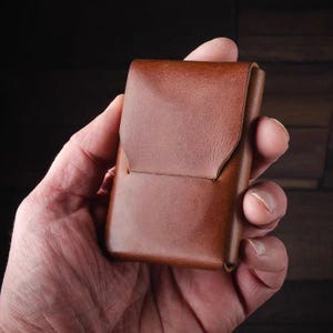 May include: A brown leather wallet with a flap closure. The wallet is held in a hand, showcasing its compact design and rich color. The leather appears to be of high quality, with a smooth texture and subtle variations in tone.