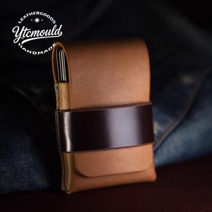 Könnte beinhalten: Handgefertigte Ledergeldbörse in Hellbraun, gesichert durch ein dunkelbraunes Band. Die Geldbörse hat eine gewölbte obere Klappe und eine kleine Vordertasche. Der Text "LEATHERGOODS Ytcmould HANDMADE" ist sichtbar.