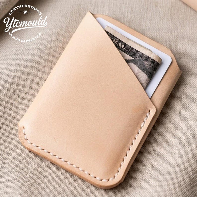 May include: A light tan leather cardholder with a folded design, stitched edges, and a white card and folded US currency visible inside. The cardholder is handmade and features the text "LEATHERGOODS Ytcmould HANDMADE".