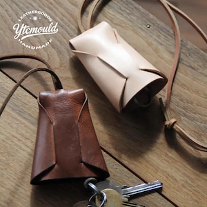 May include: Two leather key holders are shown. One is light tan, the other is brown. Both have a distinctive folded design and a leather strap. The keychains are on a wooden surface. The text "Leathergoods Ytcmould Handmade" is in a circle.