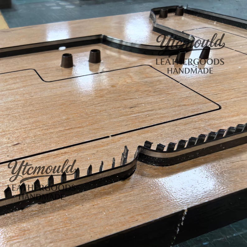 K&ouml;nnte beinhalten: Eine Holzform f&uuml;r Lederarbeiten mit schwarzen Schneidkanten und dem Text "Ytcmould LEATHERGOODS HANDMADE". Die Form weist verschiedene Formen und Aussparungen auf, darunter ein rechteckiger Bereich und mehrere kleine zylindrische Pfosten.