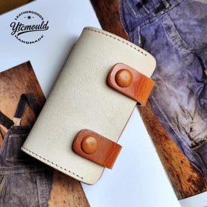 Op de afbeelding: Een crèmekleurige leren portemonnee met twee bruine leren bandjes en knoopsluitingen. De handgemaakte portemonnee heeft een gestikte rand. Het logo "LEATHERGOODS Ytcmould HANDMADE" is linksboven zichtbaar.