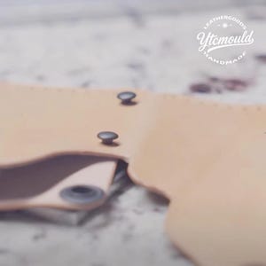 K&ouml;nnte beinhalten: Nahaufnahme eines hellbraunen Lederartikels mit zwei dunklen Metallnieten. Der Artikel scheint handgefertigt zu sein, mit einem sichtbaren gerollten Rand. Das Logo "Leathergoods Ytcmould Handmade" befindet sich in der oberen rechten Ecke.