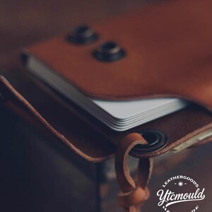 K&ouml;nnte beinhalten: Nahaufnahme einer handgefertigten braunen Ledergeldb&ouml;rse mit einem Stapel wei&szlig;er Karten. Die Geldb&ouml;rse hat zwei Metallnieten und einen Lederriemen mit einem Metallring. Der Text "LEATHERGOODS Ytcmould HANDMADE" ist sichtbar.