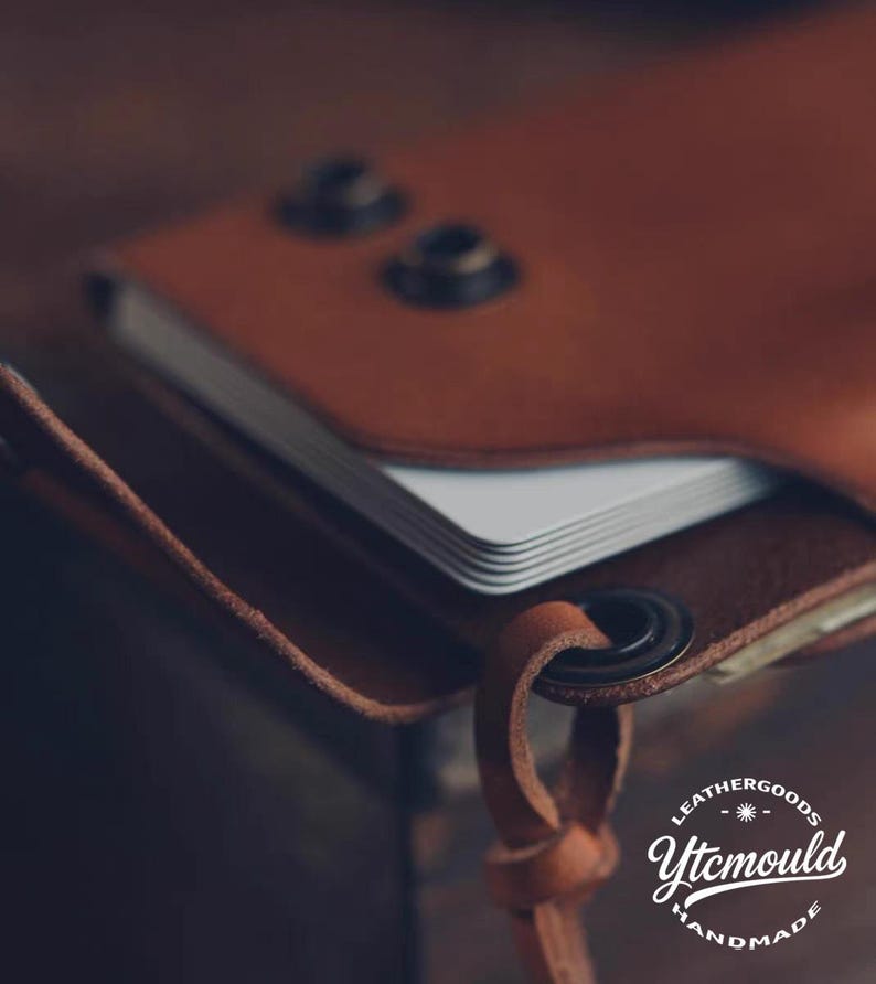 K&ouml;nnte beinhalten: Nahaufnahme einer handgefertigten braunen Ledergeldb&ouml;rse mit einem Stapel wei&szlig;er Karten. Die Geldb&ouml;rse hat zwei Metallnieten und einen Lederriemen mit einem Metallring. Der Text "LEATHERGOODS Ytcmould HANDMADE" ist sichtbar.