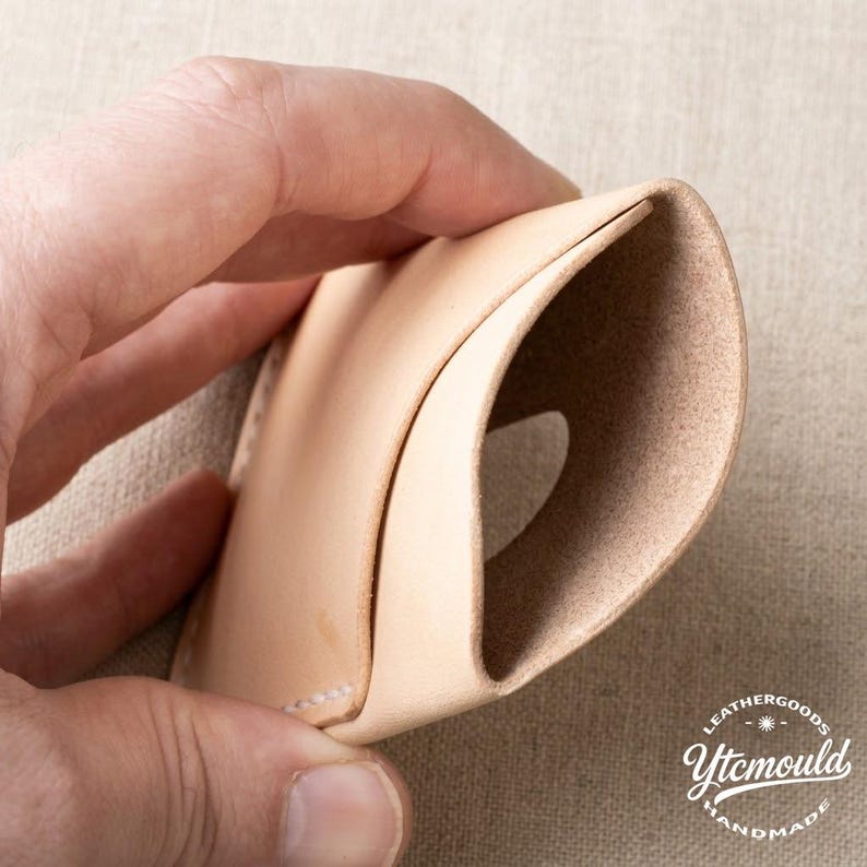 May include: A light beige leather cardholder, held open to display the interior. The handmade wallet is made of smooth, natural-colored leather. The stitching is visible along the edge. The logo "LEATHERGOODS Ytcmould HANDMADE" is in the bottom right corner.