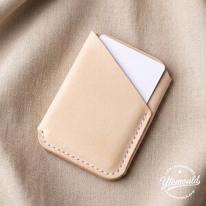 May include: A light tan leather card holder with a white card partially visible inside. The card holder has a stitched border and a minimalist design. The background is a neutral tan fabric. The card holder is a handmade item.