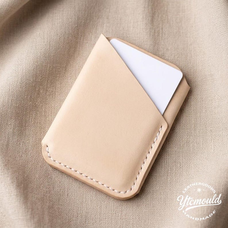 May include: A light tan leather card holder with a white card partially visible inside. The card holder has a stitched border and a minimalist design. The background is a neutral tan fabric. The card holder is a handmade item.