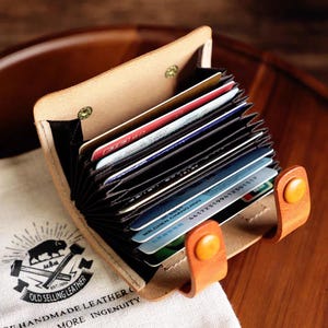 Op de afbeelding: Open beige leren kaarthouder met meerdere creditcards. De portemonnee heeft oranje knoopsluitingen. Een canvas tas met de tekst "OLD SELLING LEATHER" staat op de achtergrond.