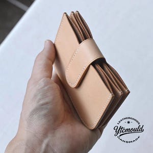 May include: A light tan leather wallet with a flap closure and a strap. The wallet is held in a hand, showcasing its compact design. The stitching is visible, and the wallet appears to be handmade. The logo "LEATHERGOODS Ytcmould HANDMADE" is visible.
