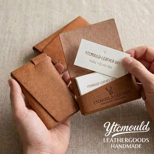 Può includere: Portacarte di credito in pelle marrone fatti a mano. Un portacarte è aperto, mostrando biglietti da visita bianchi con il testo "YTCMOULD-LEATHER ART" e un numero di telefono. Il marchio "Ytcmould" è visibile.
