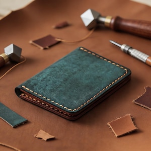 May include: A handcrafted leather wallet in teal and brown, with visible stitching. The wallet is surrounded by leather scraps and tools, suggesting a handmade product. The stitching is a contrasting light brown color.