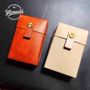Puede incluir: Dos estuches rectangulares de cuero, uno en naranja vibrante y otro en beige claro. Cada estuche tiene una solapa asegurada con un botón dorado. Los estuches están hechos a mano, como indica el texto "LEATHERGOODS Ytcmould HANDMADE" en un diseño circular.