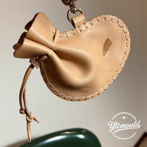 May include: A light tan leather pouch with a drawstring closure and a gold-coloured clasp. The pouch has a stitched border and a small logo. The words "Leathergoods Ytcmould Handmade" are in the bottom right corner.