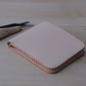 May include: A light beige leather wallet with white stitching. The wallet is square and folded, with visible layers. Crafting tools are in the background, suggesting handmade craftsmanship. The wallet is a neutral color, suitable for various styles.