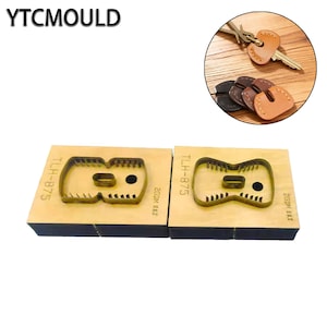 Puede incluir: Dos moldes de madera para artesanía en cuero con un diseño de llavero. Los moldes son rectangulares con una forma de llavero recortada. El texto "YTCMOULD" está en la parte superior. Una llave y llaveros de cuero están en la esquina superior derecha.