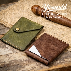 Può includere: Due porta carte in pelle. Uno è verde oliva con patta e chiusura a scatto in ottone. L'altro è marrone, aperto e mostra una carta bianca. Il testo "Ytcmould LEATHERGOODS HANDMADE" è visibile.