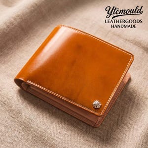 Könnte beinhalten: Ein braunes Lederportemonnaie mit weißen Nähten und einem silberfarbenen Zierelement. Das Portemonnaie ist rechteckig und hat eine glänzende Oberfläche. Der Text "Ytcmould LEATHERGOODS HANDMADE" ist sichtbar.