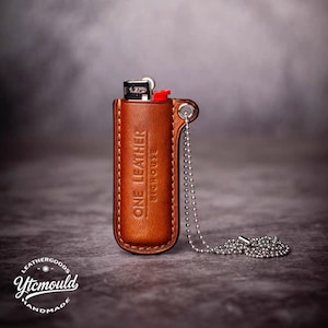 Op de afbeelding: Een bruine leren aanstekerhouder met de tekst "ONE LEATHER BICHOUSE" in reliëf op de voorkant. De houder heeft een zilveren ketting en bevat een zwart-zilveren aansteker met een rode ontstekingsknop. De achtergrond is een vage grijstint.