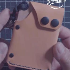 K&ouml;nnte beinhalten: Ein handgefertigter, hellbrauner Lederkartenhalter mit schwarzen N&auml;hten und schwarzen Druckkn&ouml;pfen. Der Kartenhalter tr&auml;gt den Text "LEATHERGOODS Ytcmould HANDMADE" in wei&szlig;er Schrift. Der Kartenhalter wird von einer Person in den H&auml;nden gehalten.