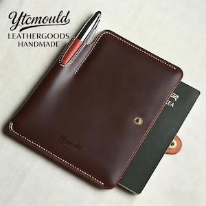 May include: A brown leather notebook cover with white stitching and a pen holder. The cover has the text "Ytcmould LEATHERGOODS HANDMADE". A pen is inserted in the holder, and a dark green notebook is partially visible.
