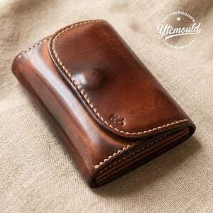 May include: A handcrafted brown leather wallet with a curved flap and white stitching. The wallet has a small embossed design and the text "LEATHERGOODS Ytcmould HANDMADE".
