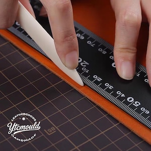 May include: A close-up of a craftsperson using a bone folder and a ruler to work with orange leather. The ruler is black with white markings. The text "Ytcmould HANDMADE LEATHERGOODS" is visible in the lower left corner.