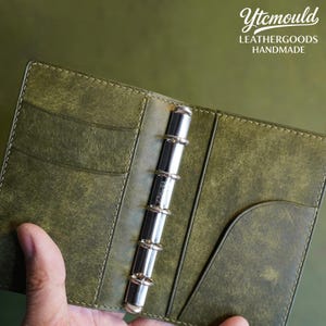 May include: Open, olive green leather binder with a silver metal ring mechanism. The binder has pockets and stitching details. The text "Ytcmould LEATHERGOODS HANDMADE" is visible in the upper right corner.