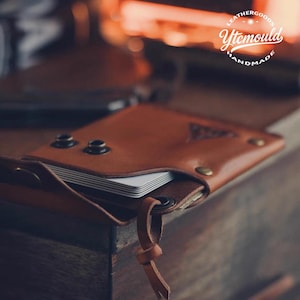 May include: A brown leather wallet with a flap closure, holding a stack of white cards. The wallet has metal rivets and a leather strap. The text "Leathergoods Ytemould Handmade" is visible.