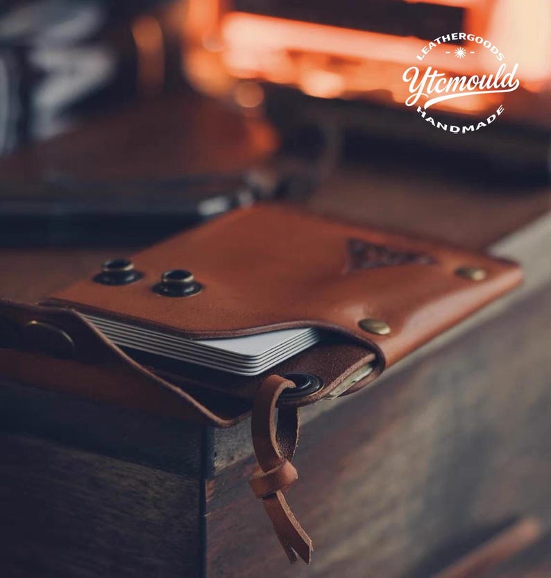 K&ouml;nnte beinhalten: Ein braunes Lederportemonnaie mit Klappenverschluss, das einen Stapel wei&szlig;er Karten enth&auml;lt. Das Portemonnaie hat Metallnieten und einen Lederriemen. Der Text "Leathergoods Ytemould Handmade" ist sichtbar.