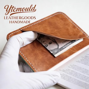 May include: A brown leather wallet with white stitching, holding a five-dollar bill. The wallet is open, revealing a glimpse of a book. The text "Ytcmould LEATHERGOODS HANDMADE" is visible.