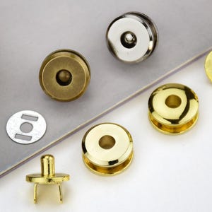 Può includere: Una collezione di chiusure magnetiche circolari in metallo con varie finiture, tra cui ottone anticato, argento lucido e oro brillante. Il set include componenti maschio e femmina, alcuni con rebbi e rondelle piatte di supporto in metallo.