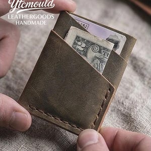 Może przedstawiać: Ręcznie robiony, brązowy skórzany portfel z widocznymi szwami. Portfel jest otwarty, ukazując banknot i kartę. Tekst "Ytcmould LEATHERGOODS HANDMADE" jest widoczny w lewym górnym rogu.