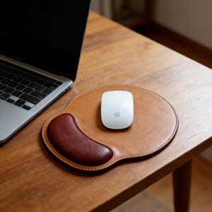 Puede incluir: Un mouse pad de cuero marrón con un reposamuñecas burdeos y un ratón de ordenador blanco. El mouse pad está sobre un escritorio de madera junto a un ordenador portátil.
