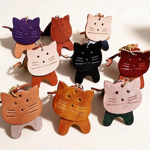 May include: A selection of leather cat-shaped keyrings in diverse colours, such as purple, brown, black, and pink. Each keyring features a smiling cat face with stitched details and a metal ring for attachment. The keyrings are designed with small arms and legs.