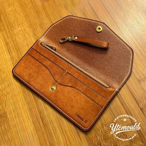 May include: Open, tan leather wallet with a matching wrist strap. The wallet has multiple card slots and a zippered pocket. The text "Lemon II" is visible on the front. The wallet is on a wooden surface.