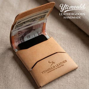 Op de afbeelding: Een lichtbruine leren portemonnee, open om contant geld en een zwarte kaart te laten zien. De portemonnee heeft een minimalistisch ontwerp met een klepsluiting. De tekst "Ytc mould Leathergoods Handmade" is zichtbaar.