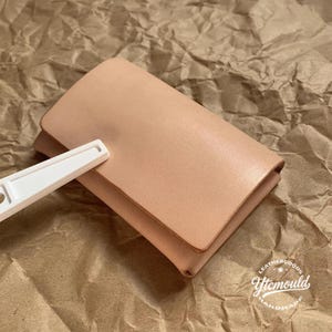 Puede incluir: Una cartera de cuero de color beige claro con cierre de solapa. Una herramienta blanca descansa sobre la cartera. La cartera está sobre un fondo de papel marrón. La cartera es un producto hecho a mano.