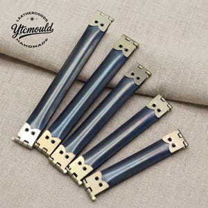 May include: Five blue-grey leather bag handles with silver-colored metal accents. The handles are arranged diagonally, showcasing their rectangular shape and decorative metal ends. The image includes the text "LEATHERGOODS Ytcmould HANDMADE".