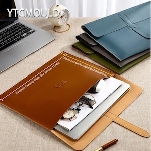 May include: A brown leather document holder with a flap closure, containing a laptop, mouse, and accessories. The text on the holder reads "Maximum Capacity Width Size 335mm/13.98in" and "Maximum Capacity Height Size 245mm/9.65in". Several other document holders in blue, gray, and green are stacked in the background.