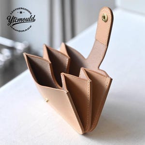 Może przedstawiać: Jasnobrązowy skórzany portfel z wieloma otwartymi przegródkami. Posiada zapięcie na zatrzask i uchwyt. Tekst "LEATHERGOODS Ytcmould HANDMADE" jest wydrukowany w lewym górnym rogu. Ręcznie robiony.